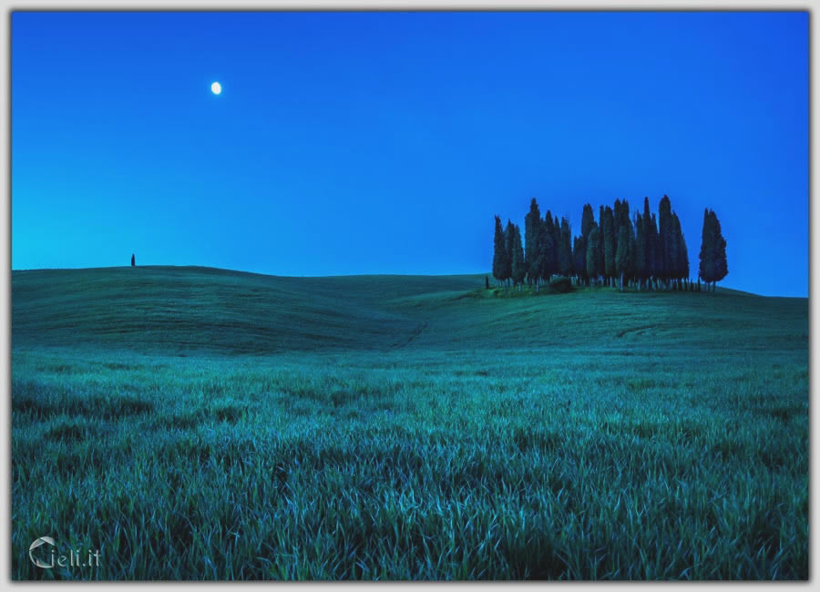 Luna luminosa e chiara nel cielo azzurro intenso, con pace intorno e colline nitide. Immensità avvolta dalla pace e ombre immobili.